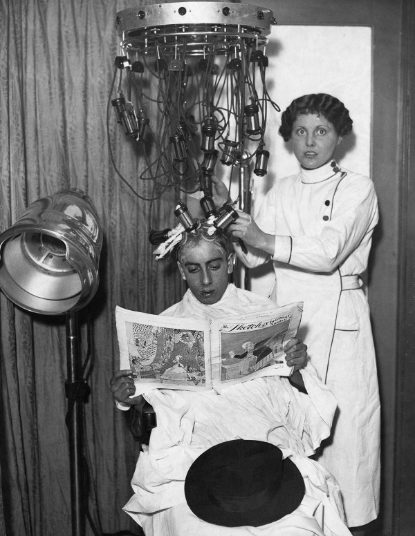 ca. 1930s, UK --- A beautician in a white coat attaches the tubes and pads of a hair treatment machine to a man's scalp as he reads a paper contentedly. Probably England, ca. 1930s. --- Image by © Hulton-Deutsch Collection/CORBIS